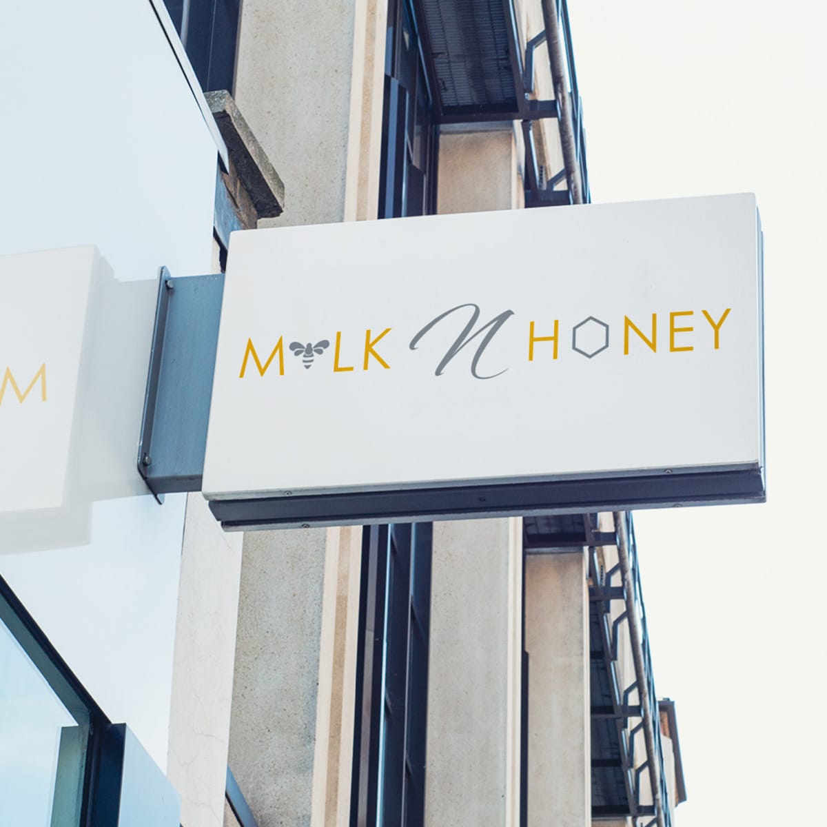 White storefront sign displaying the Milk n Honey logo in yellow, gray, and white against an architectural background