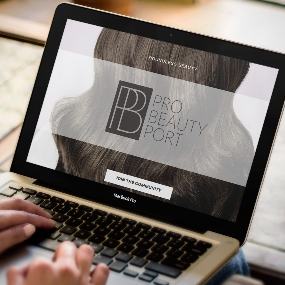 Omaha Logo Designer for Hire A laptop screen on a wooden desk shows a homepage layout with a monogram logo centered over a full-bleed photo of voluminous brown hair and a pale button below, a website mockup designed by an Omaha logo designer.