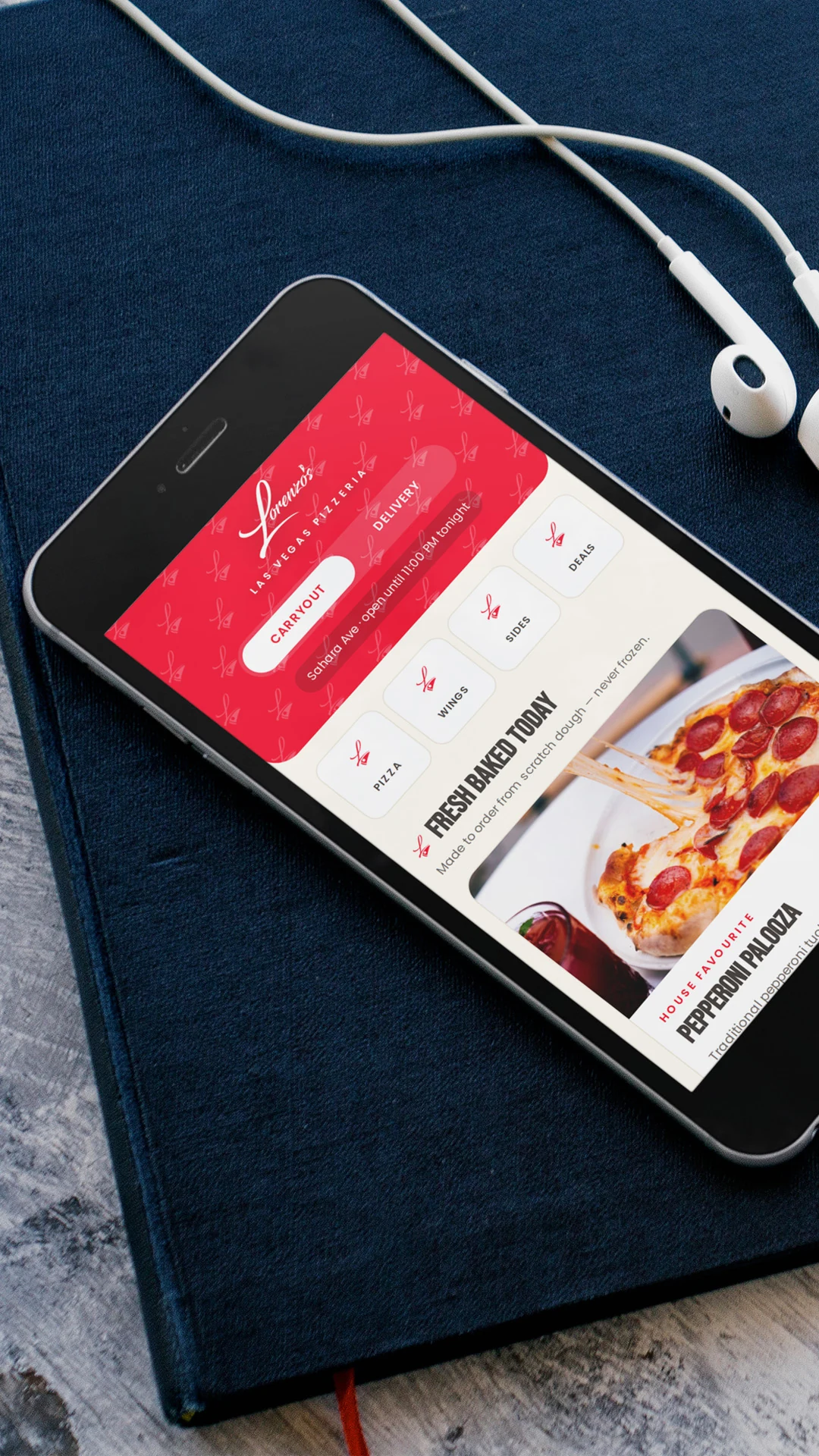 pizza-restaurant-branding-phone-mockup Phone on a navy notebook showing Lorenzo's pizza ordering homepage beside white earbuds.