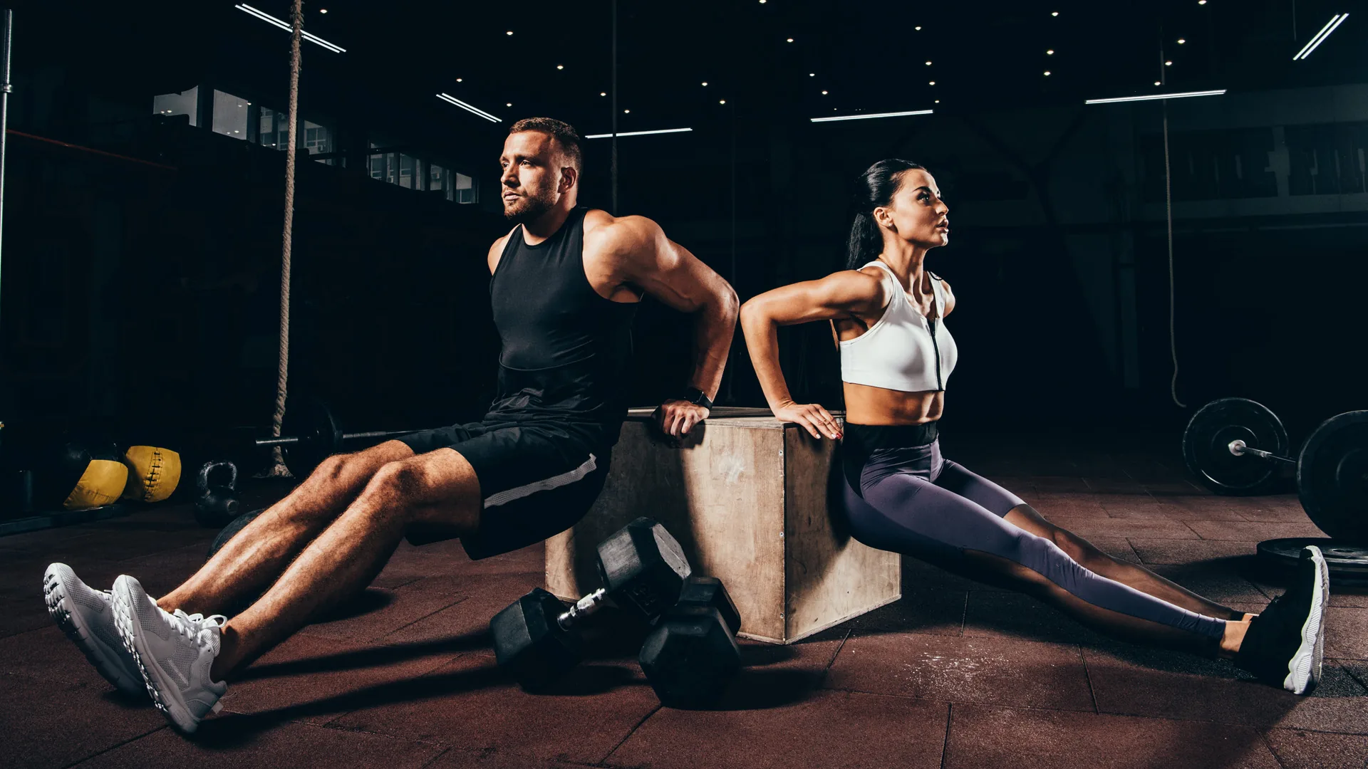 Couple gym workout industry design Man and woman doing tricep dips on a wooden plyo box in a dark gym.