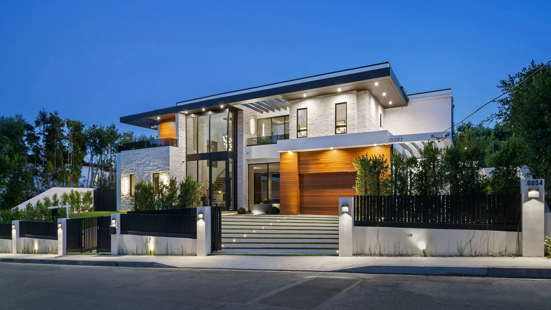 Modern luxury home Modern two-story house with wood and stone accents, lit up at dusk behind a fence.