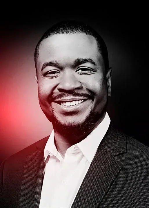website-designer-kenal-louis-headshot Smiling man in a dark blazer and white shirt against a black-and-red backdrop, Kenal Louis, website designer.